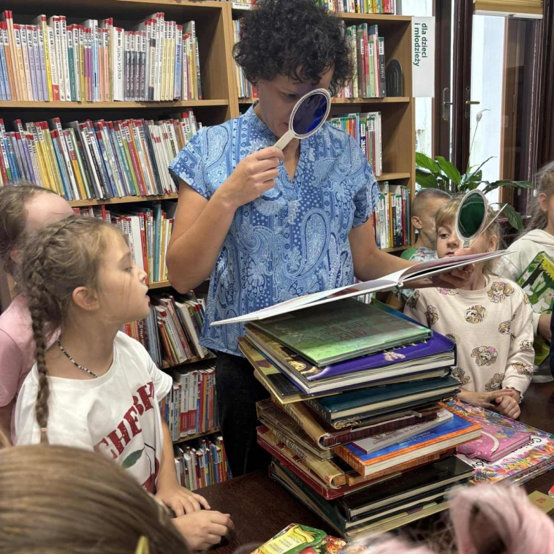 Pani patrzy przez lupę na książki, uczniowie patrzą na książkę